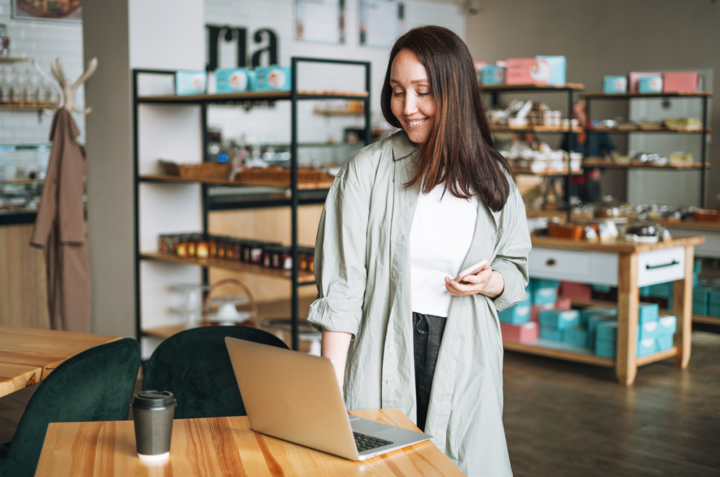 Adult brunette business woman practicing how to increase customer retention rates in casual working with laptop using mobile in cafe